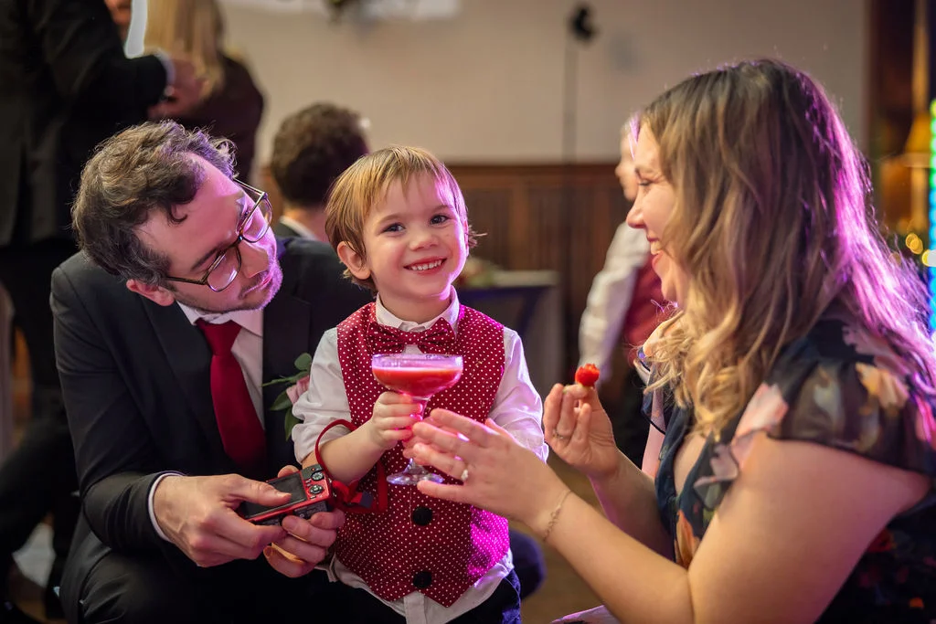 A young pageboy in a polka dot waistcoat smiles happily while holding a red cocktail glass, surrounded by guests at a wedding reception.