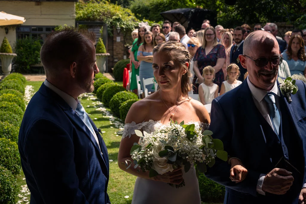 A smiling bride walks down a sunlit garden aisle holding her father's arm while looking towards the waiting groom in a blue suit.