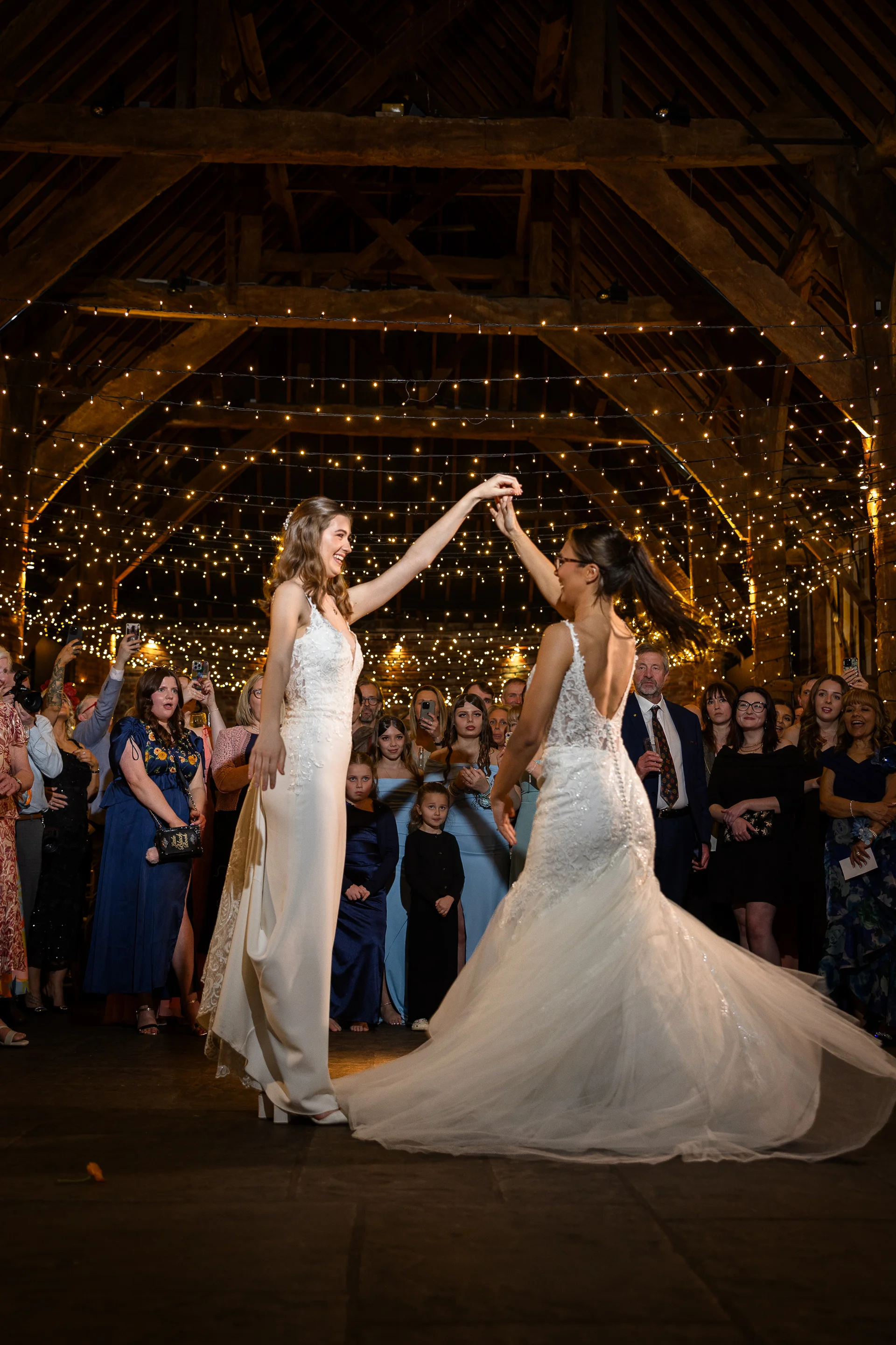 Two brides share their first dance under a stunning canopy of fairy lights inside a rustic oak framed barn surrounded by cheering guests.