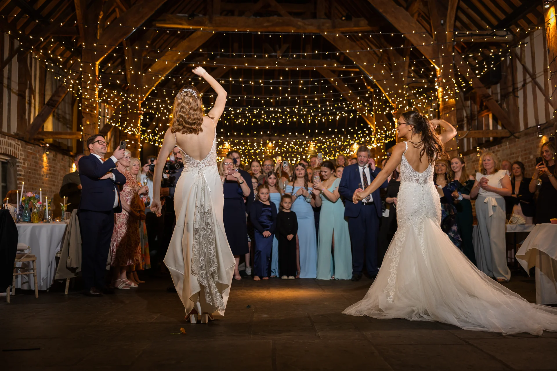 Two brides in stunning wedding dresses perform their first dance inside a rustic oak-framed barn illuminated by a magical fairy light canopy.