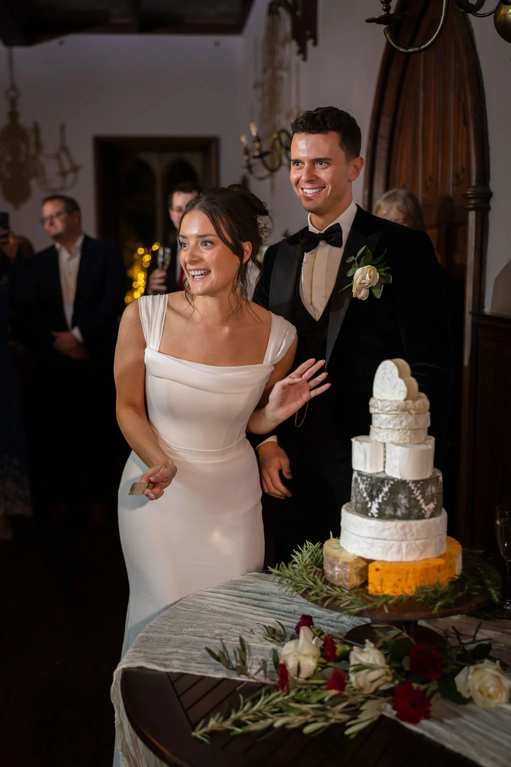 A smiling bride in a white satin gown and groom in a black tuxedo prepare to cut a multi-tiered cheese wedding cake.