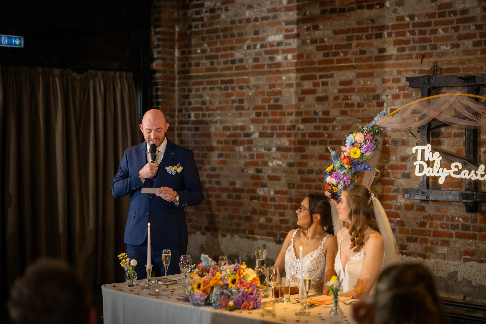 A groom in a navy suit delivers a speech using a microphone while two brides in white wedding dresses look on from the top table in an industrial brick venue.
