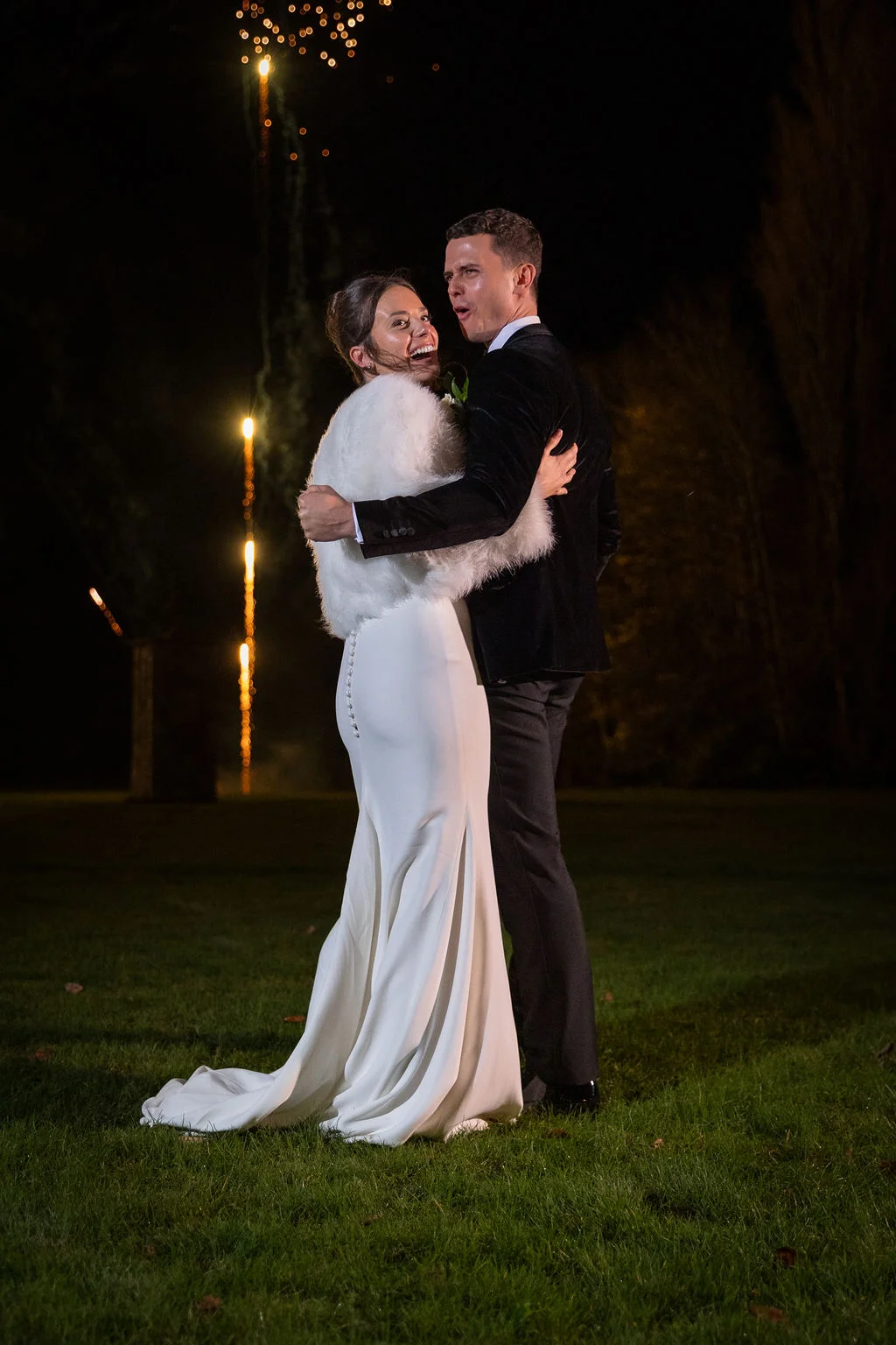 A bride in a white crepe gown with a faux fur shrug and a groom in a black tuxedo share a joyful embrace outdoors at night with distant fireworks.