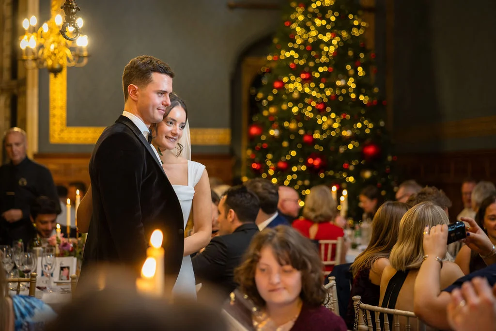 A newlywed couple stands smiling during their winter wedding reception, beautifully framed against a glowing Christmas tree and candlelit tables.