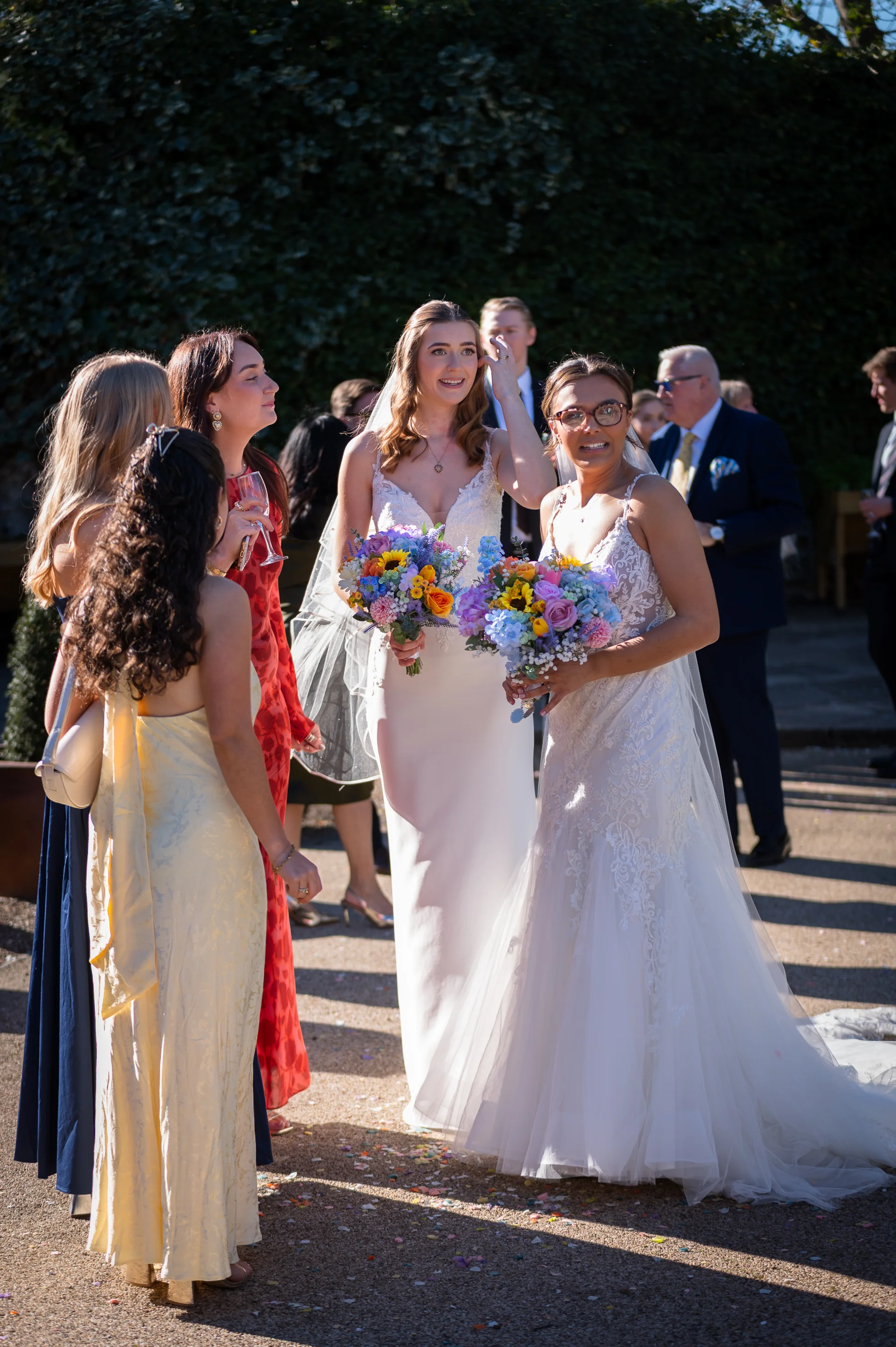 Two smiling brides in white wedding dresses holding colourful bouquets stand outdoors in bright sunshine, surrounded by wedding guests in a sunlit courtyard.