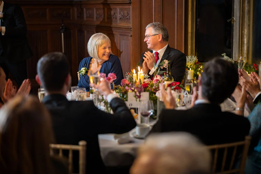 A smiling couple sits at a candlelit top table in a wood-panelled room while wedding guests applaud during the speeches.