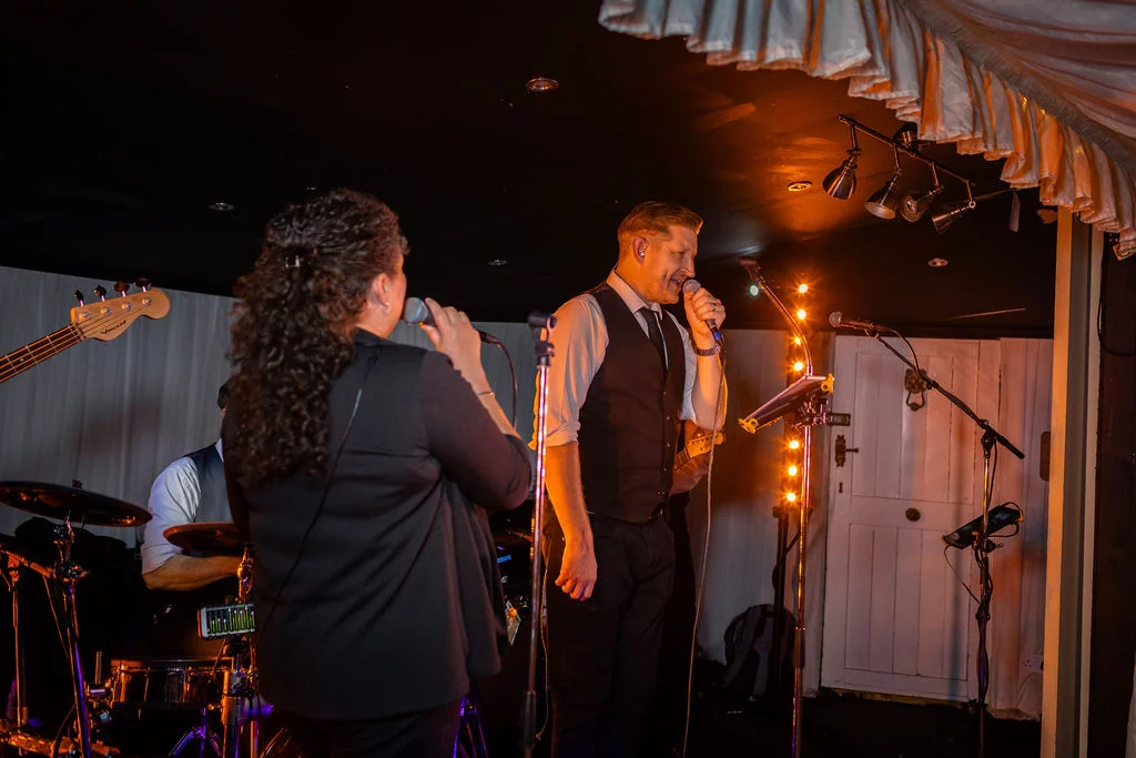 A live band performs on a stage with warm stage lighting during an evening wedding reception inside a marquee.