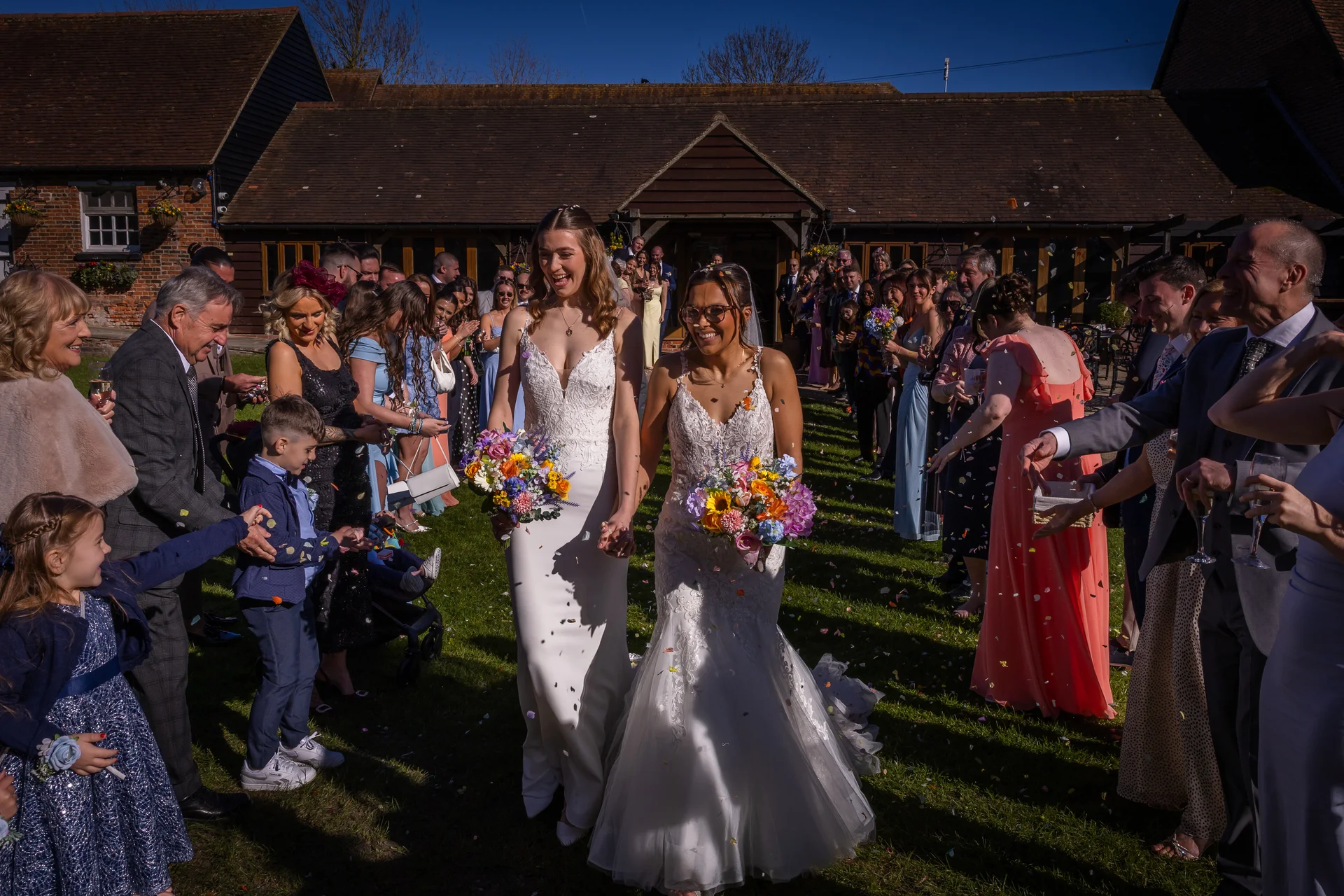 Two smiling brides in white wedding dresses walk hand in hand through a cheering crowd of guests throwing confetti outside a rustic barn venue on a sunny day.