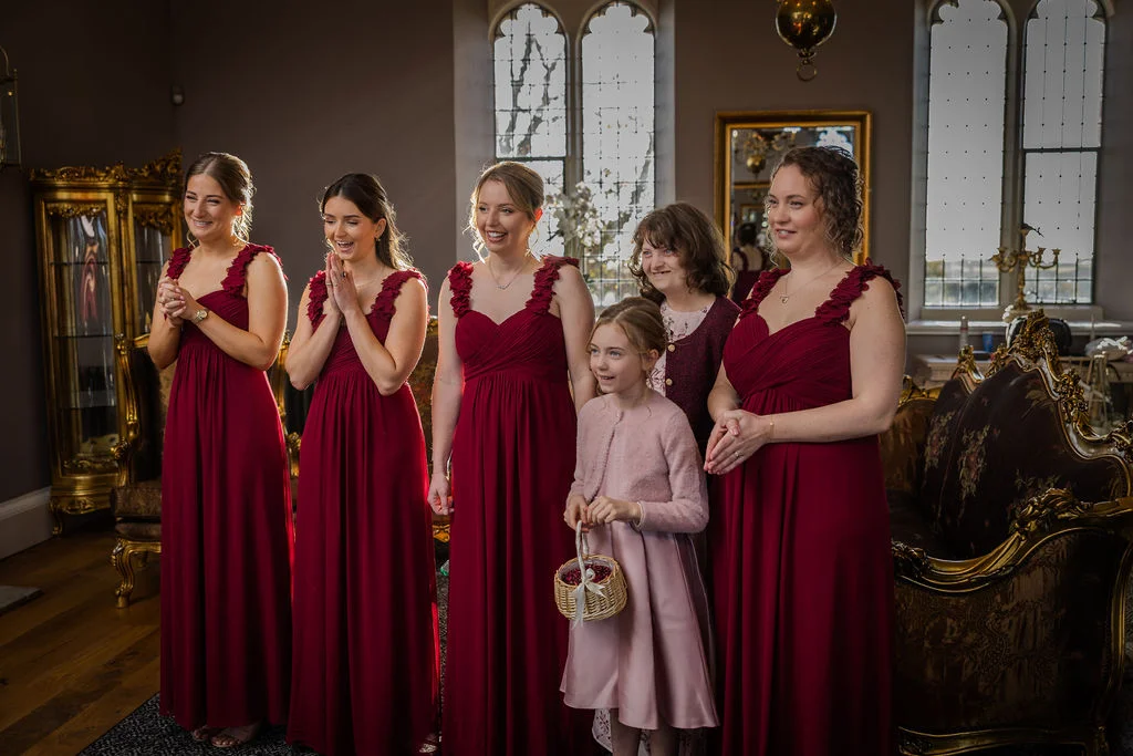 A group of bridesmaids wearing matching burgundy gowns and a flower girl holding a basket stand indoors near a tall window, smiling and laughing.