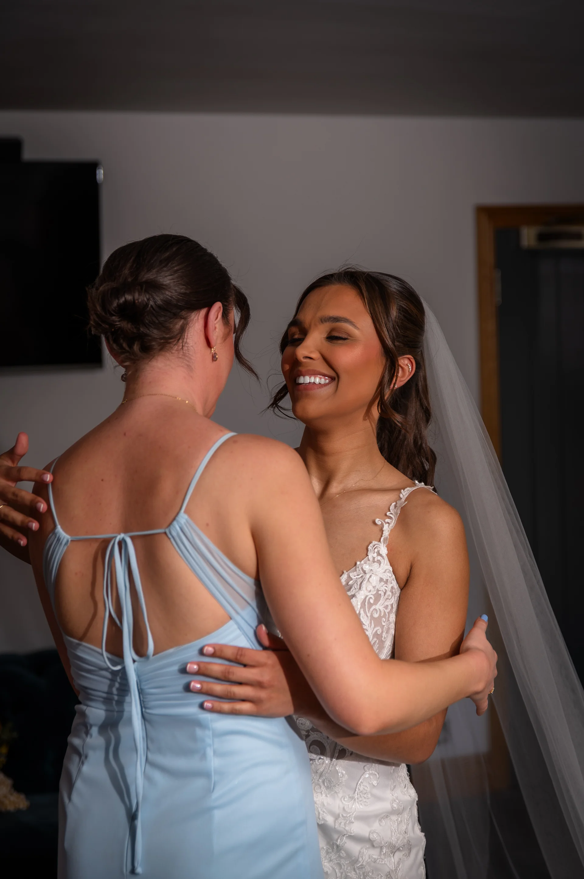 A joyful bride in a lace wedding dress smiles warmly while embracing a bridesmaid wearing a pale blue dress during wedding morning preparations.