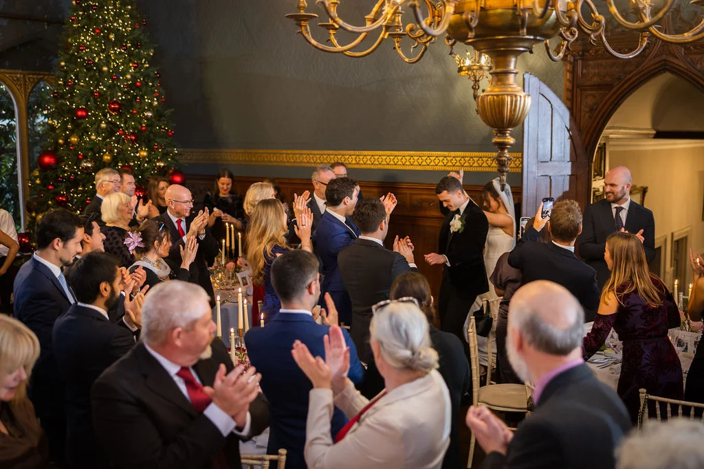 Newlyweds enter their festive Christmas wedding reception to rapturous applause from seated guests gathered around candlelit tables near a large decorated tree.