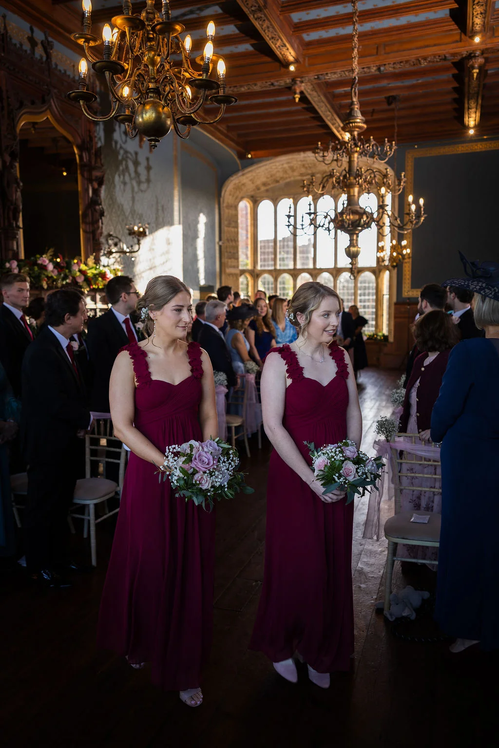 Two bridesmaids in matching burgundy dresses and holding floral bouquets walk down the aisle in a grand historic hall.