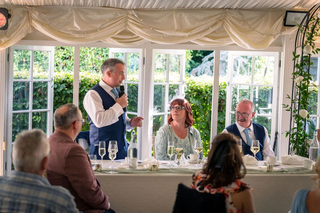 A groom delivers a speech holding a microphone at the head table during a bright conservatory wedding reception.