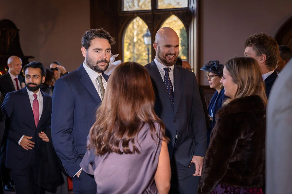 A group of smartly dressed wedding guests mingle and laugh together inside an elegant country house venue with arched windows.