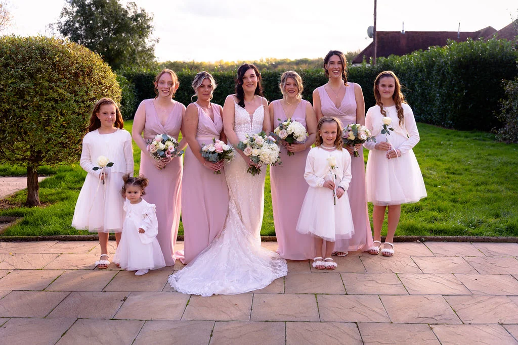 The bride stands in a lace gown holding a bouquet, surrounded by her bridesmaids in matching pink dresses and flower girls on a paved patio.