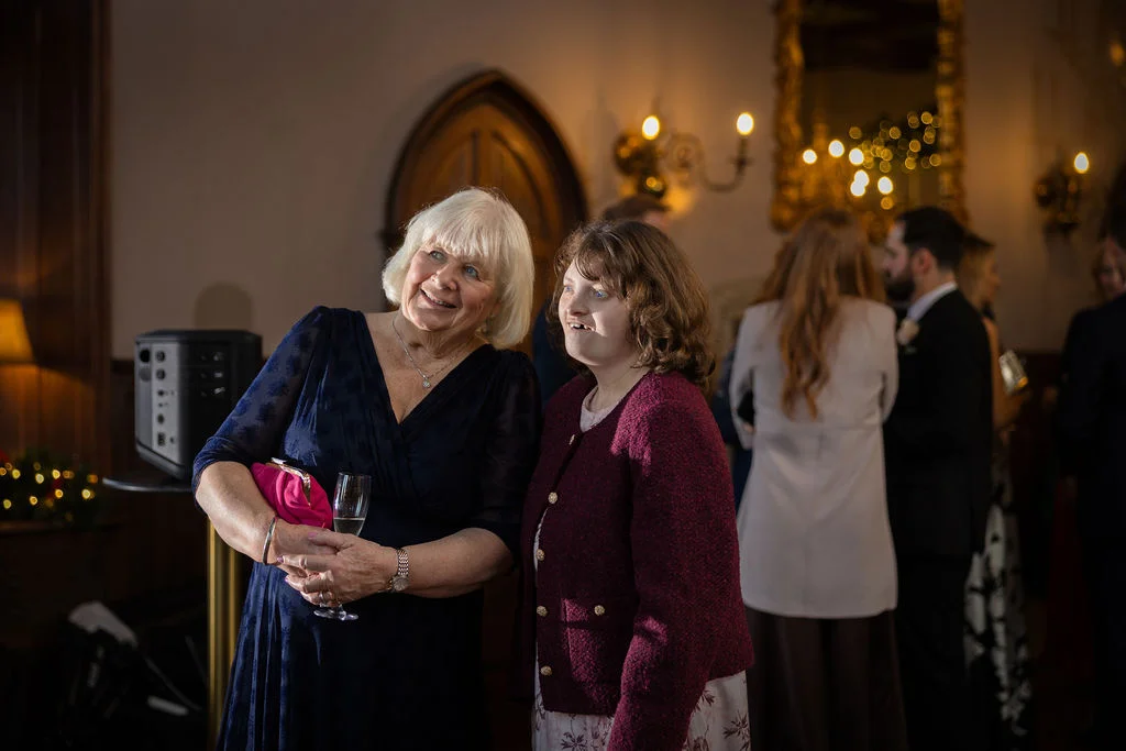 Two smiling wedding guests looking attentively towards speeches inside an elegantly lit historical venue room.