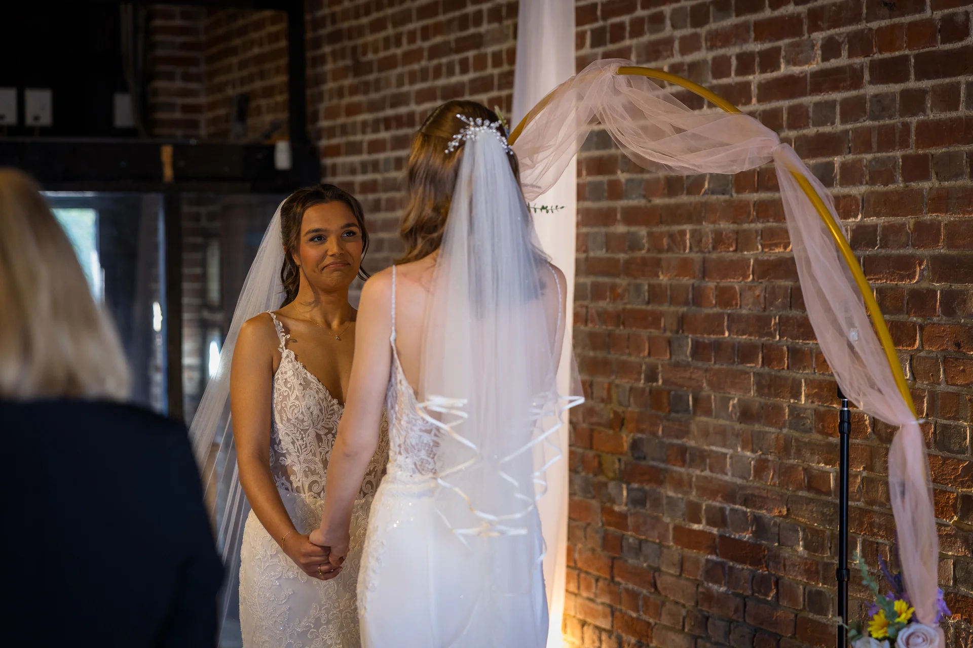 Two brides hold hands and exchange emotional vows during an indoor wedding ceremony in front of an exposed brick wall and a circular arch draped with sheer pink fabric.