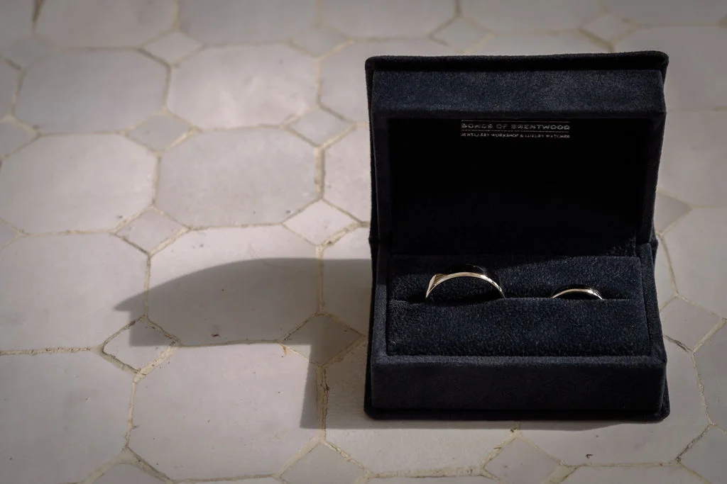 Two wedding bands sit side by side inside a dark velvet ring box resting on a tiled surface.