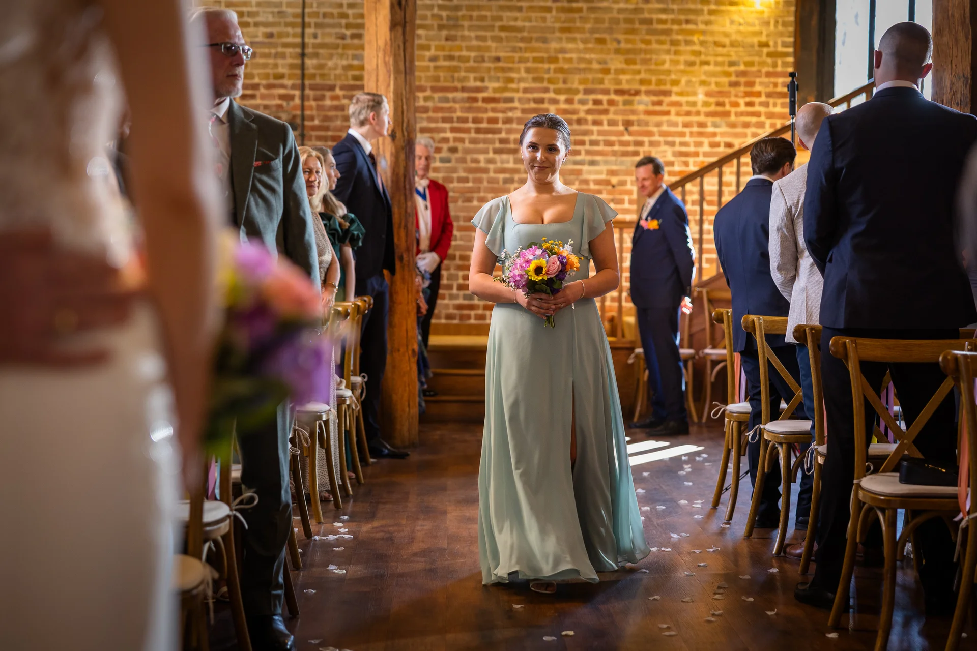 A bridesmaid wearing a sage green dress with flutter sleeves and a slit walks down the wooden aisle of a rustic brick barn holding a colourful bouquet.