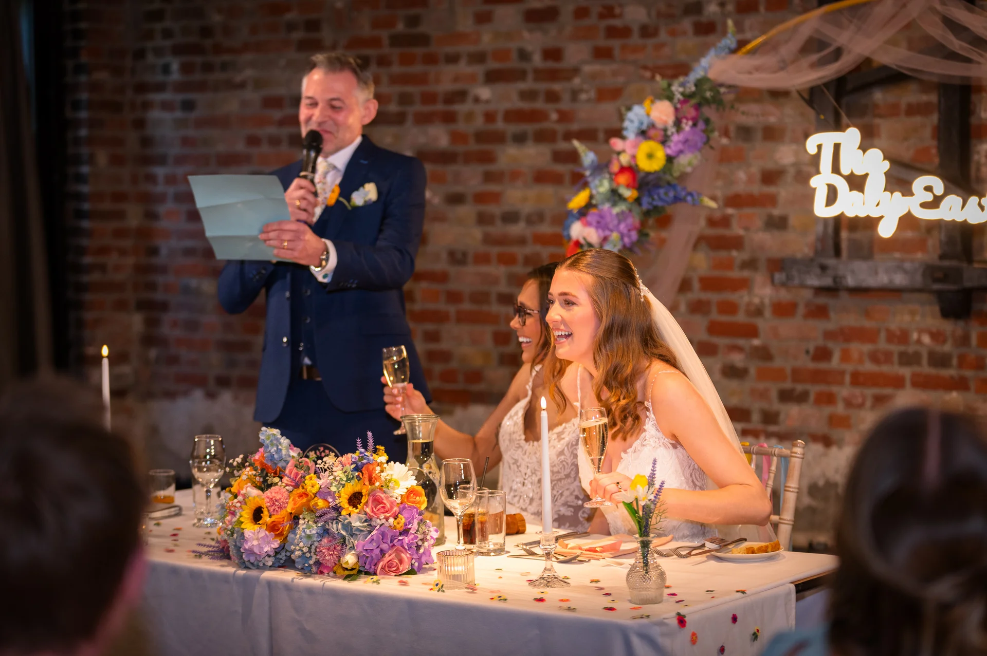 A smiling bride and a bridesmaid laugh while holding champagne glasses at the top table during the wedding speeches inside a venue with exposed brick walls.