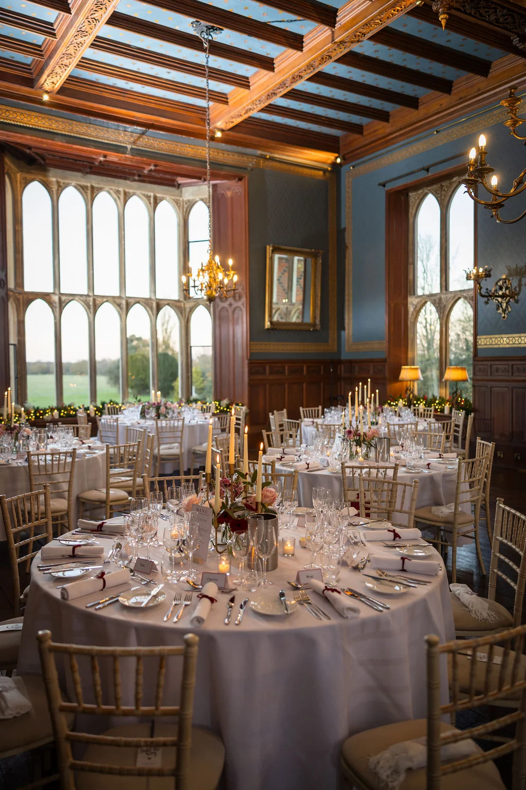 An elegant wedding reception room in a stately home features round tables set with white linens, taper candles, and floral centrepieces under a magnificent vaulted ceiling and tall arched windows.