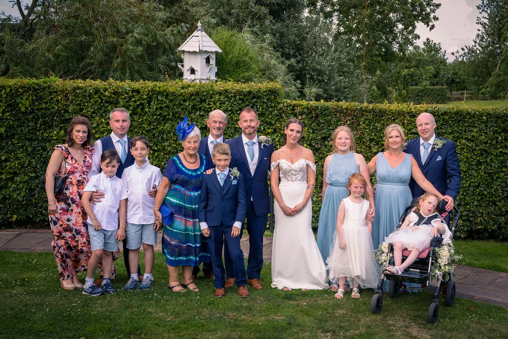 A large wedding group poses together outdoors on a lawn in front of a tall green hedge, featuring the bride in a white gown and the groom in a blue suit alongside family members and children.