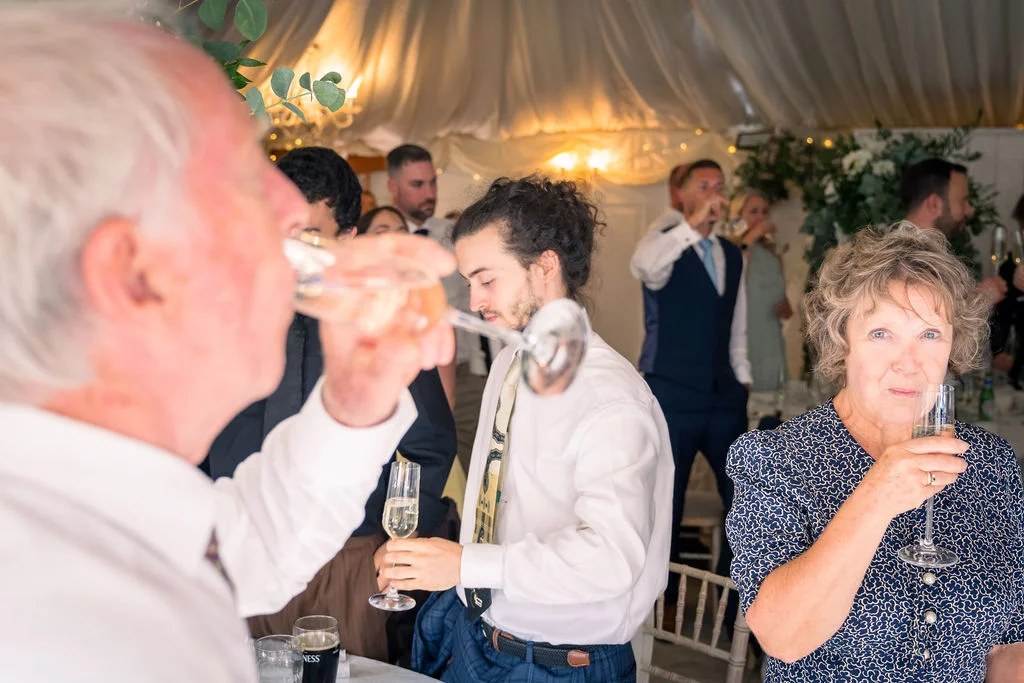 Wedding guests enjoy champagne and socialise inside a brightly lit marquee draped with soft white fabric.