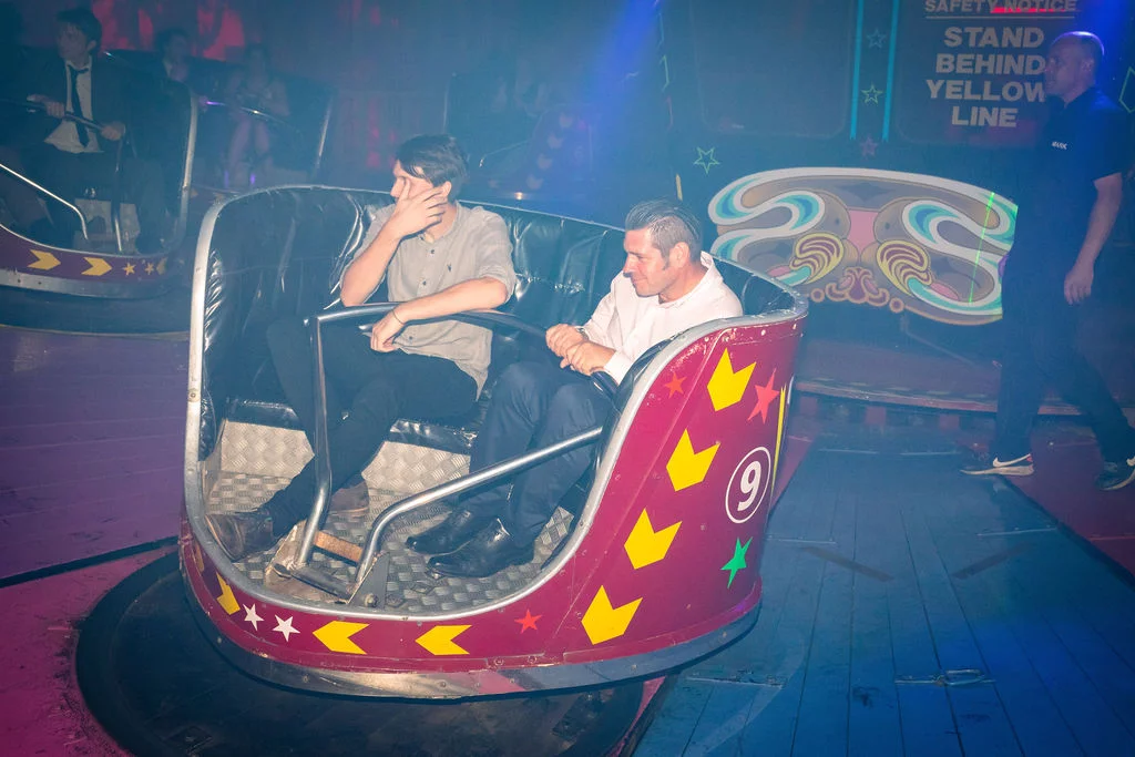 Two guests enjoy a ride on a fairground attraction during an evening wedding reception under flashing neon lights.