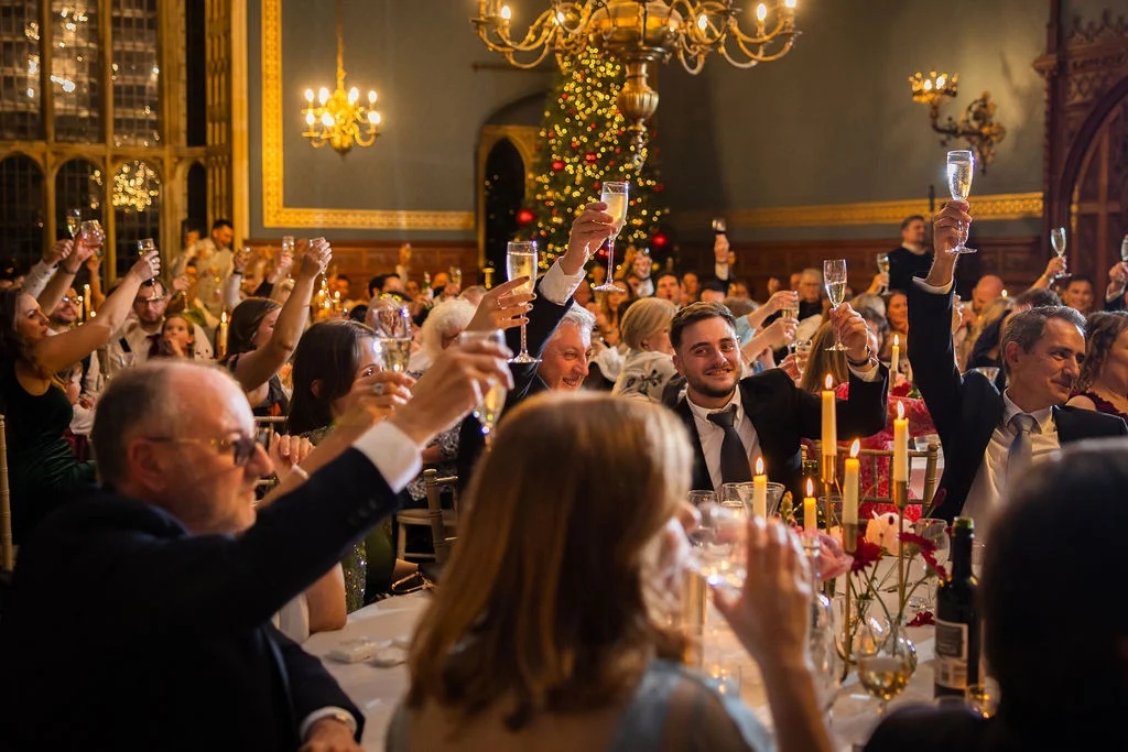Wedding guests raise their champagne glasses in a toast during a festive evening reception inside a grand candlelit ballroom adorned with a lit Christmas tree.