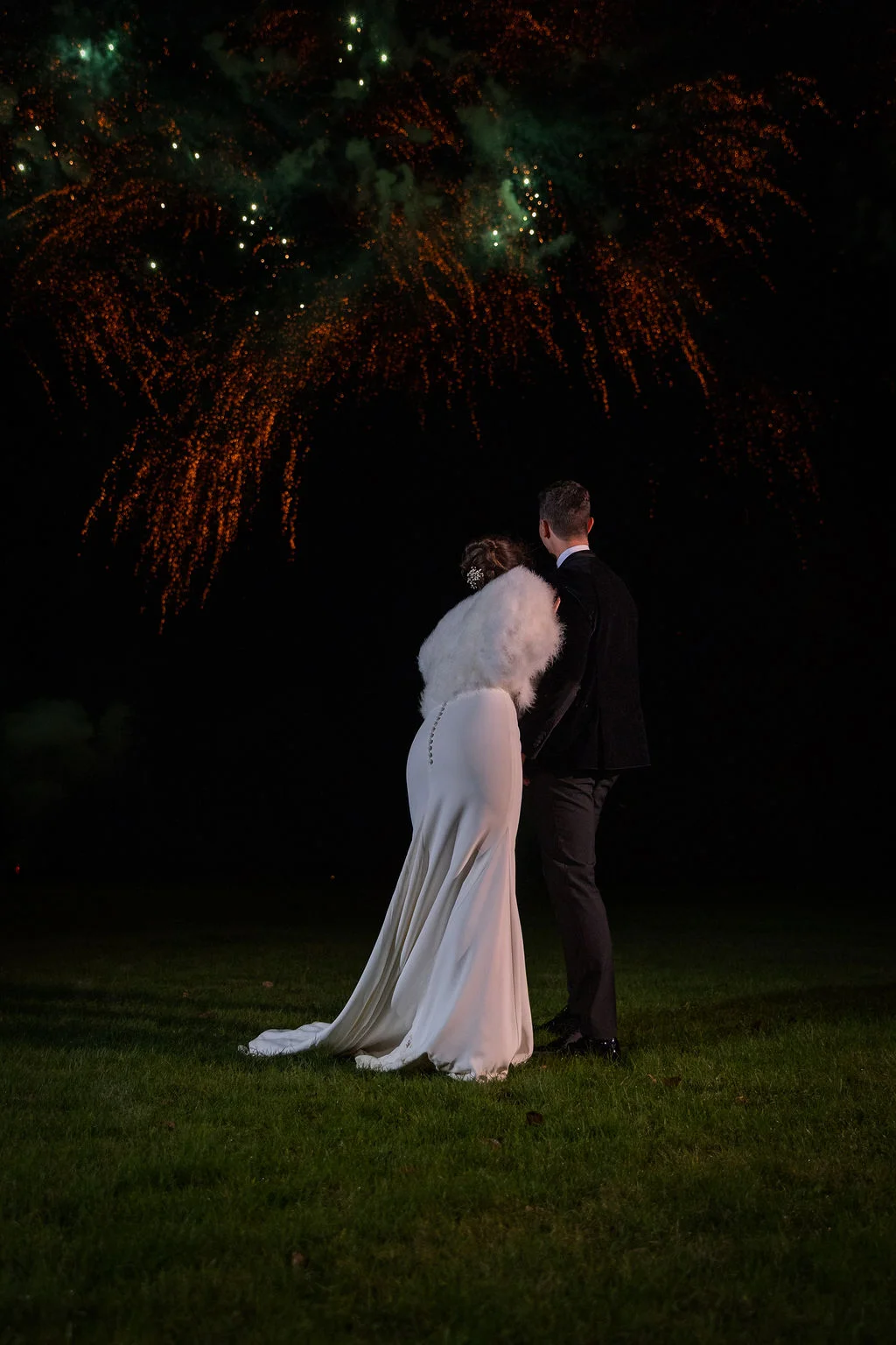 Newlyweds stand together on a lawn at night, watching a vibrant fireworks display burst across the dark sky above them.