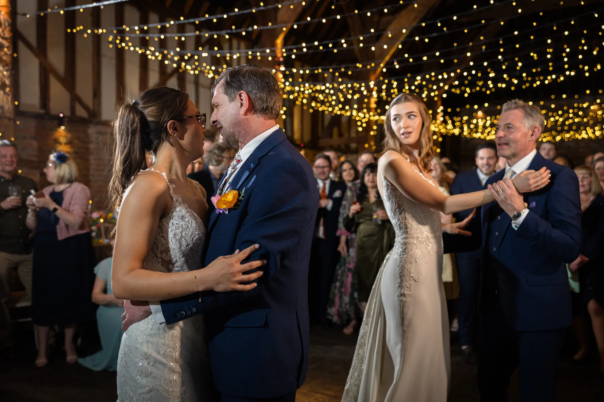 Two couples share a joyful first dance together under a dazzling canopy of fairy lights inside a rustic oak-framed barn wedding venue.