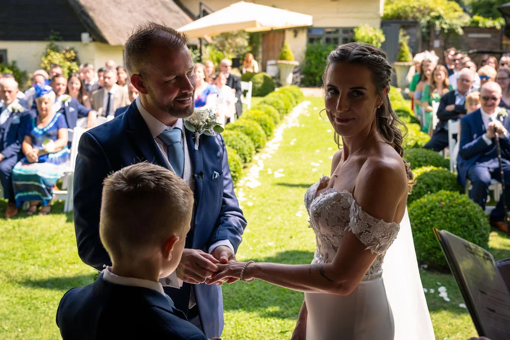 A groom gently slides a wedding ring onto the bride's finger during a sunlit outdoor garden ceremony while a young boy watches closely.