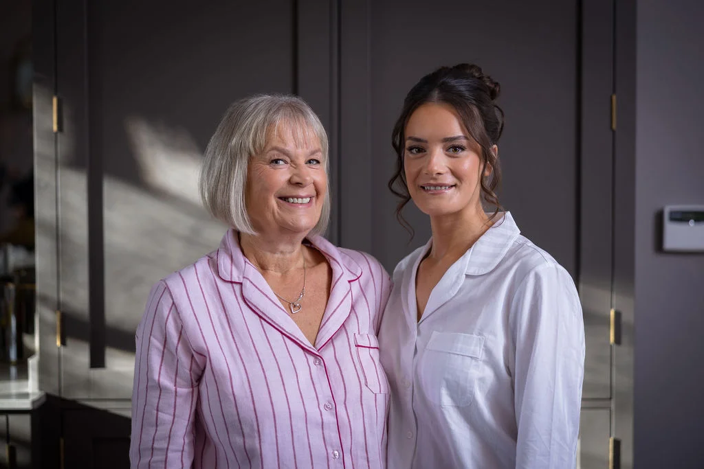 An older woman in pink striped pyjamas and a younger woman with an updo hairstyle in a white shirt smile warmly together indoors.