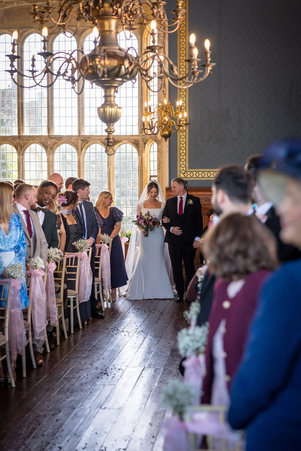 A bride walking down the aisle accompanied by her father in a grand historic hall with large windows and wooden floors.