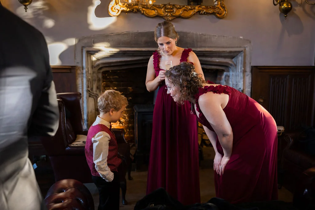Two bridesmaids in burgundy dresses lean down to chat with a young page boy wearing a red patterned waistcoat inside an atmospheric venue with a stone fireplace.