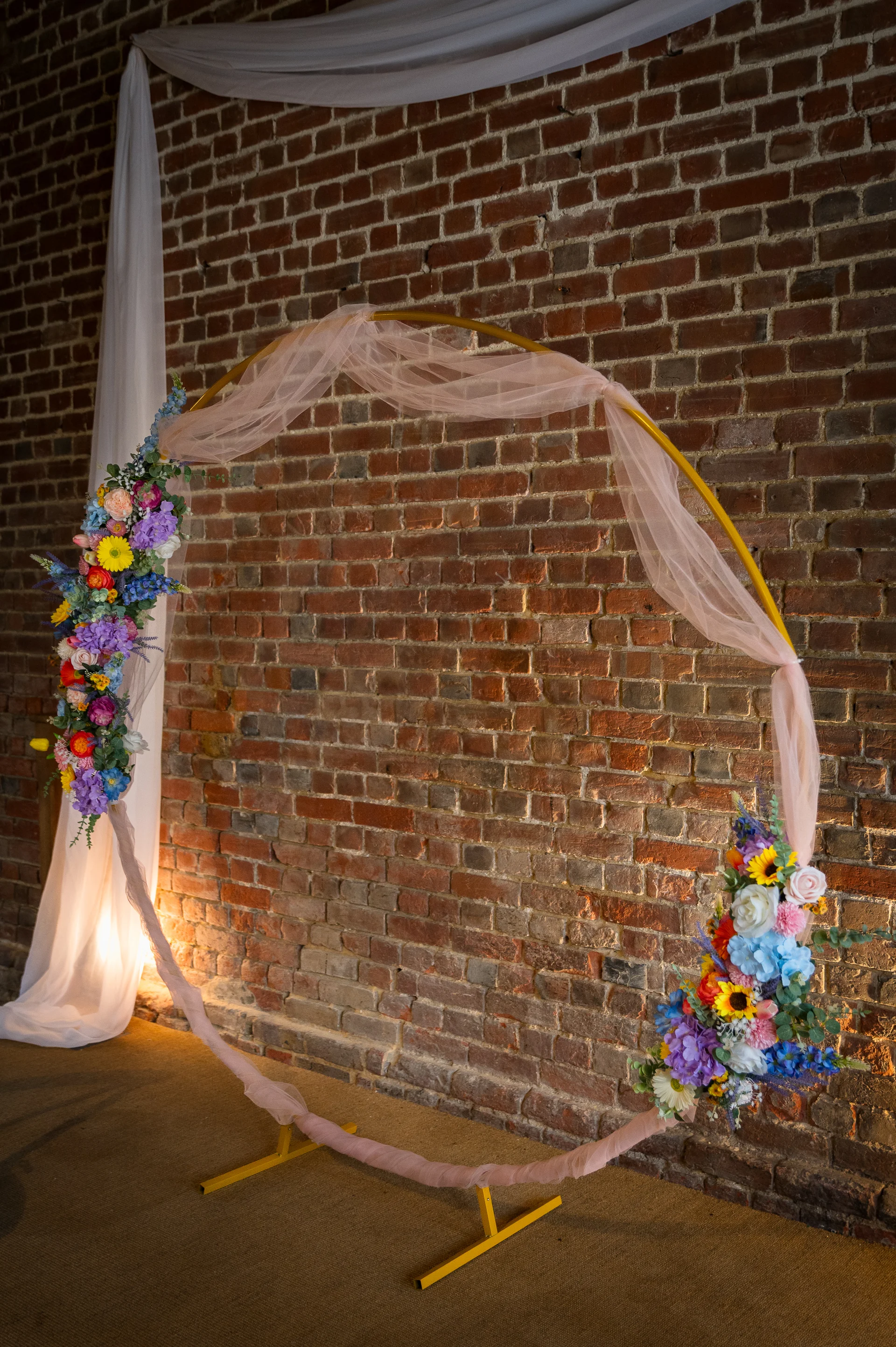 A circular gold wedding arch adorned with vibrant multi-coloured flowers and sheer pink fabric stands against an exposed brick wall in a rustic barn venue.