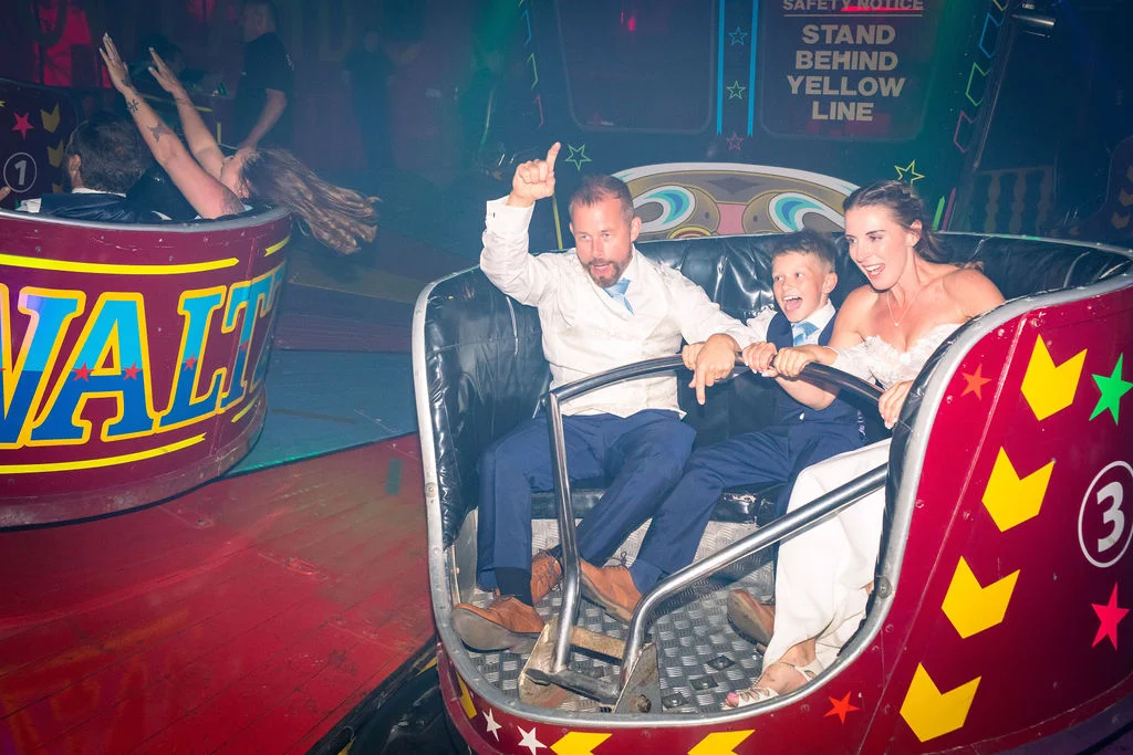 A bride, groom and young guest laugh with pure joy while riding a colorful funfair waltzer car during an energetic evening wedding reception.