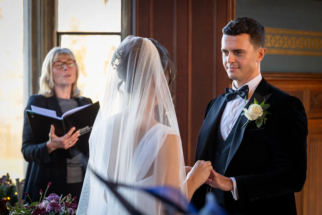 A groom in a black tuxedo and bow tie exchanges rings with his bride during an indoor ceremony, as a celebrant officiates in the background.