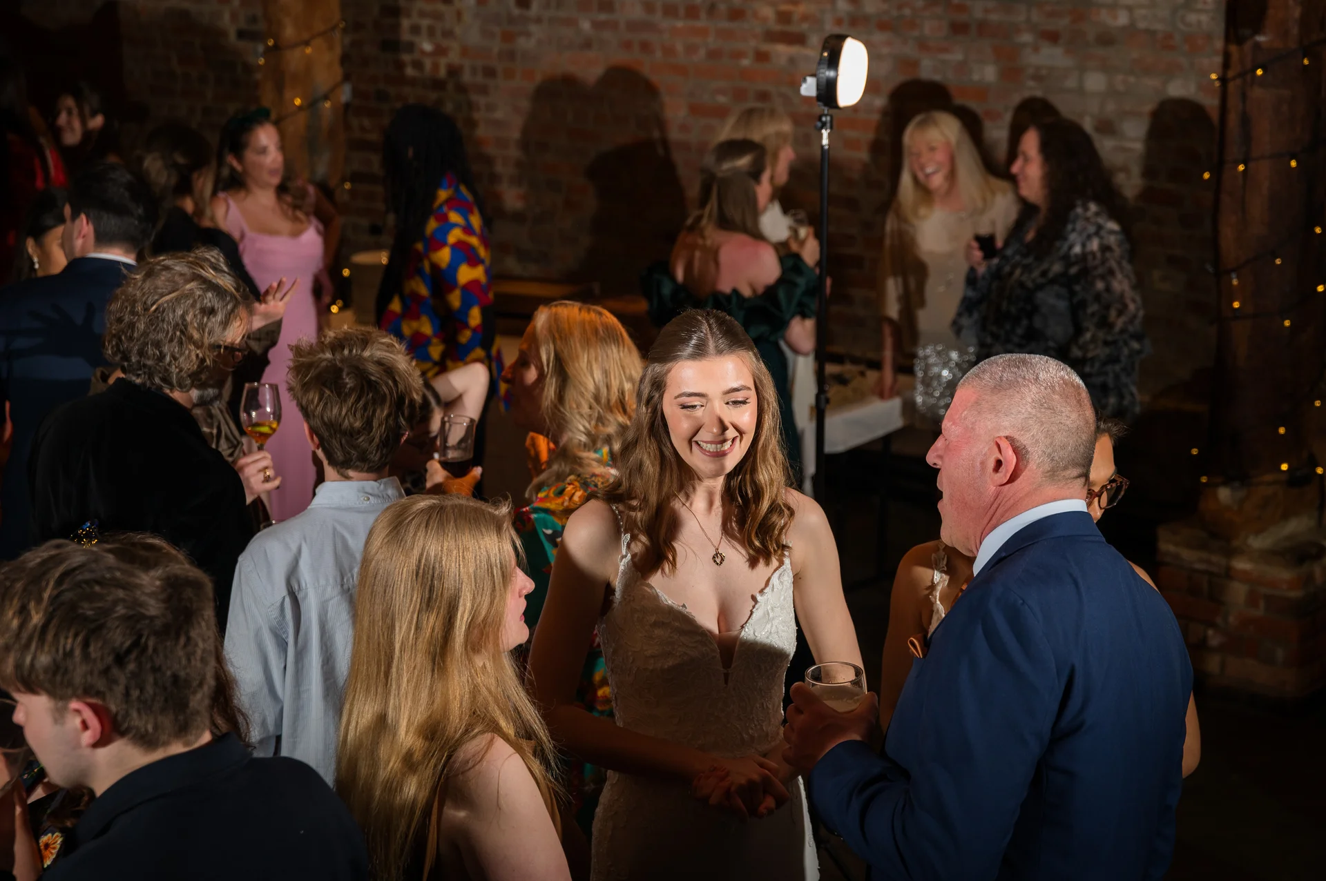 A smiling bride in a white gown chats with guests during a lively evening reception in an exposed brick venue.