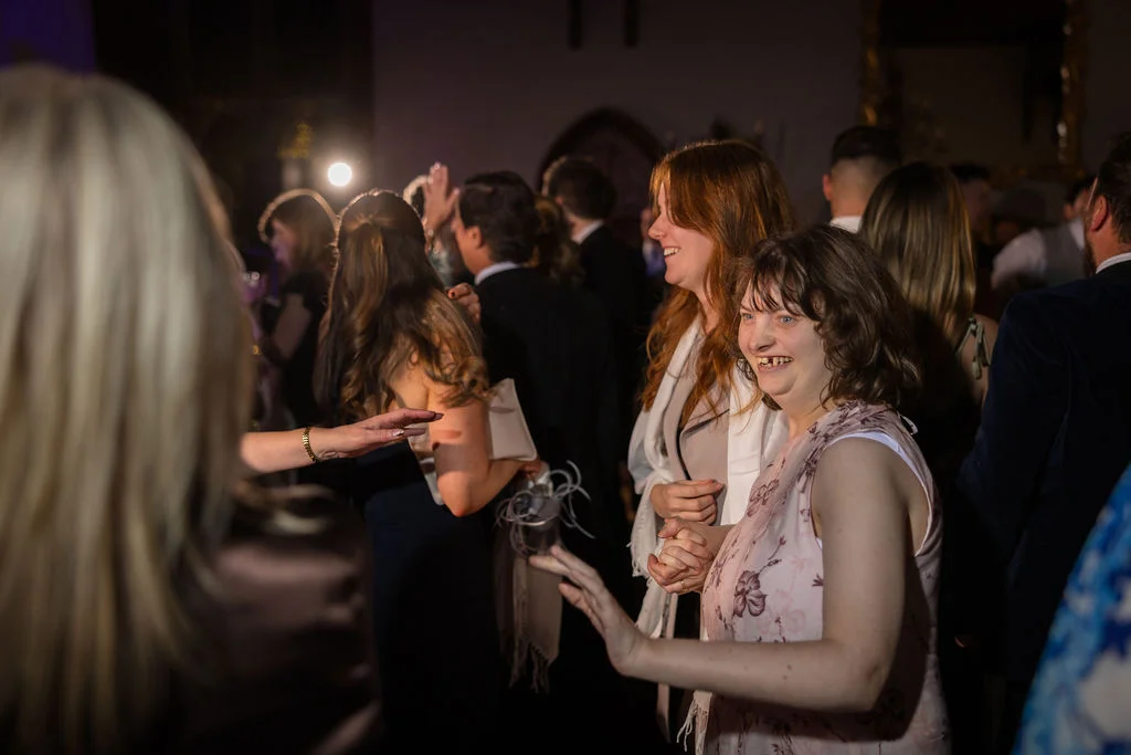 Wedding guests laugh and dance together during a lively evening reception in a dimly lit venue.
