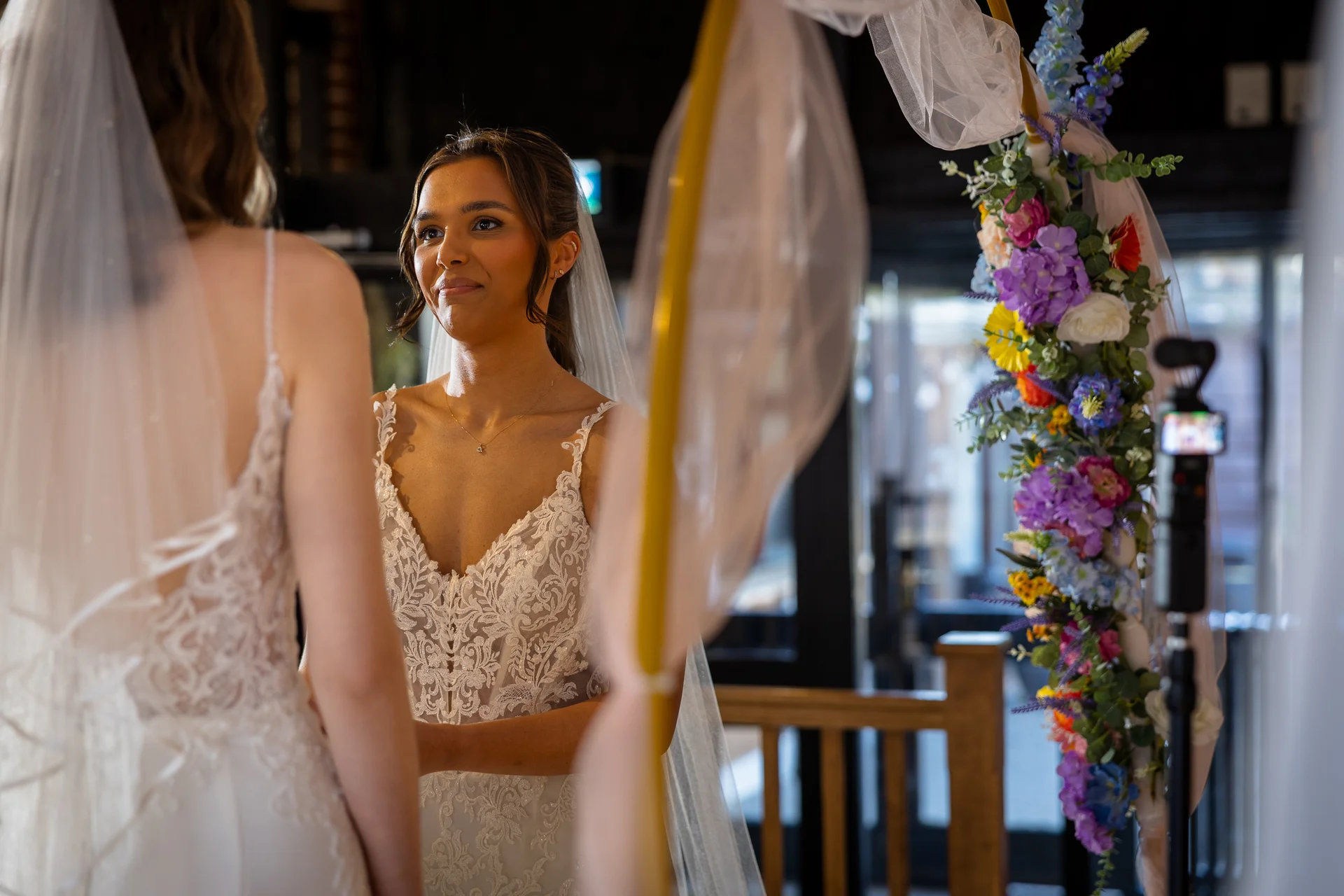 Two brides share a tender moment during their wedding ceremony beneath a vibrant floral arch adorned with colourful blooms.