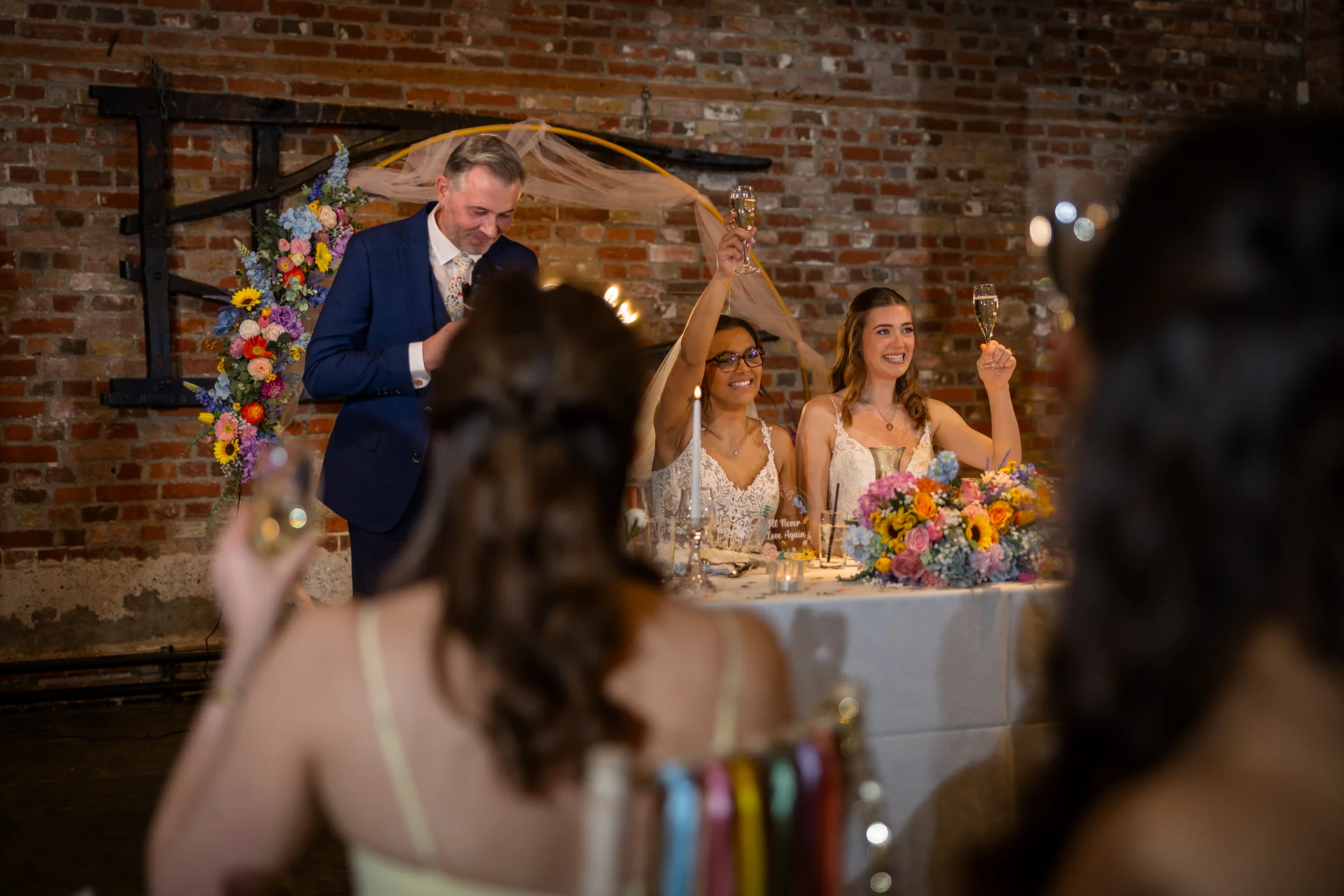 Two brides and a best man raise their champagne glasses in a toast during the wedding speeches at a rustic brick venue.