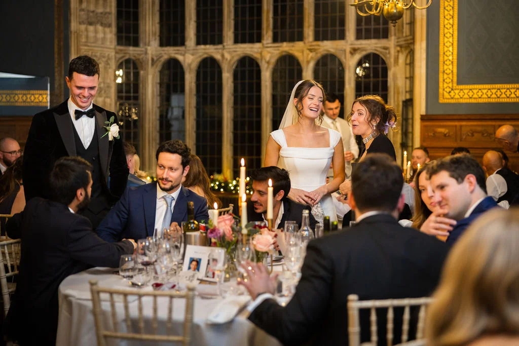 A joyful bride and guests laugh during wedding speeches in a grand hall featuring large arched windows and candlelit tables.