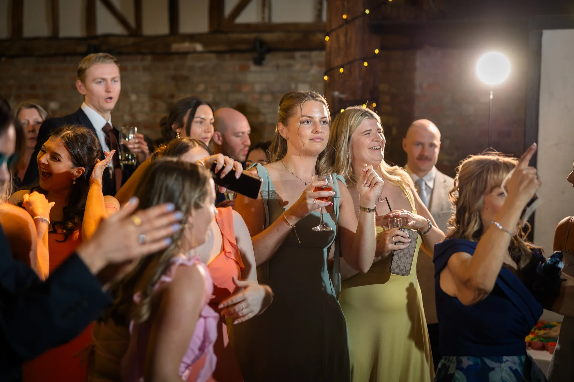 A group of wedding guests enjoying themselves during the evening reception inside a rustic barn venue with exposed brick walls.