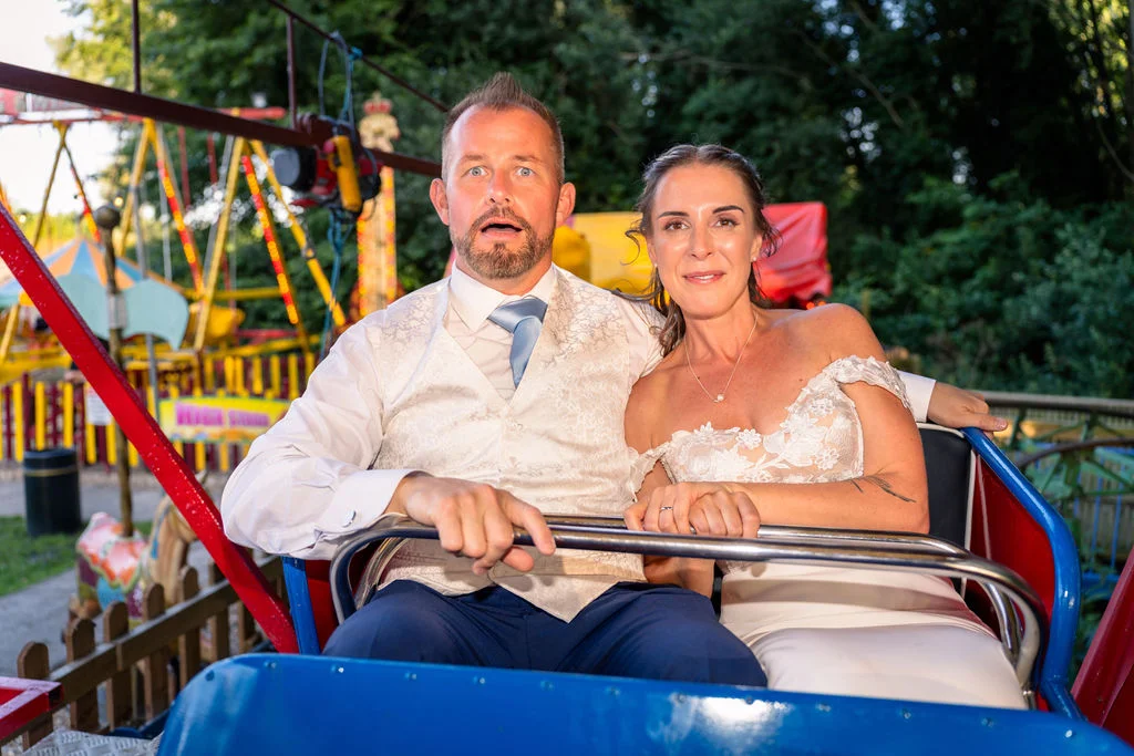 Newlyweds enjoying a playful moment together on a funfair ride, with the groom looking excitedly surprised and the bride smiling calmly in her floral lace wedding dress.