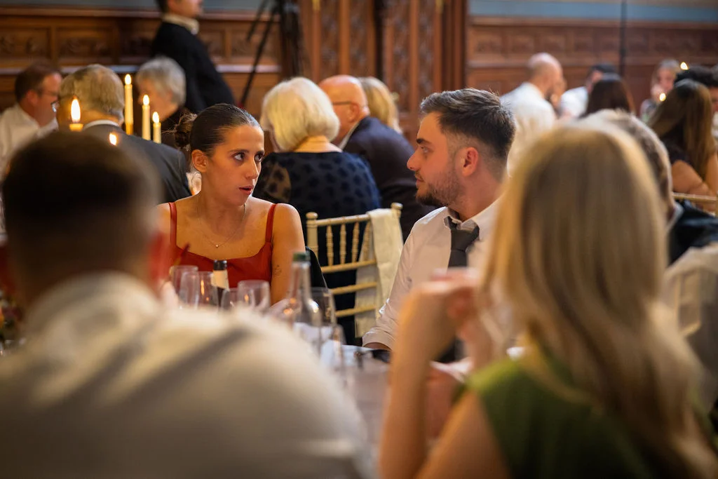 Guests enjoy lively conversation and dinner at a candlelit wedding reception seated around long banquet tables inside an elegant panelled room.