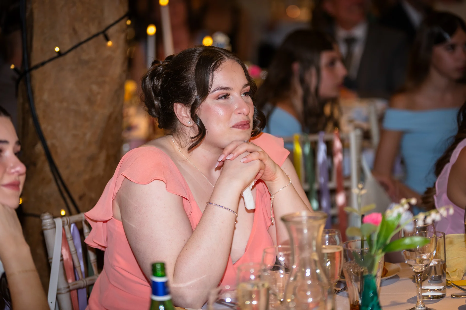 An emotional wedding guest sitting at a reception table listens intently during the speeches while holding her hands beneath her chin.