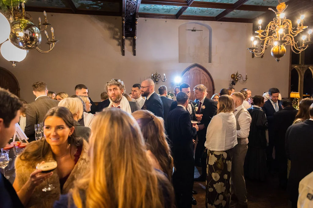 Wedding guests mingle and enjoy drinks in a grand hall featuring high ceilings, ornate chandeliers, and classic architectural details.