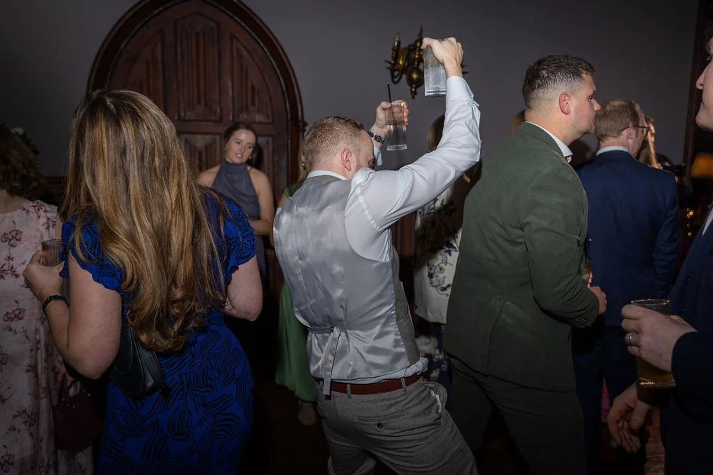 A candid shot of wedding guests dancing and socialising in a dimly lit reception venue with a large arched wooden door.