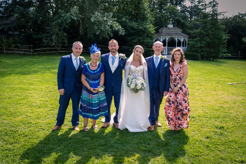 A happy wedding group stands together on a sunny lawn in front of a white gazebo, featuring the bride in a strapless gown with a chapel veil alongside the groom, parents, and guests.